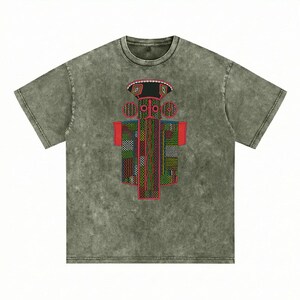 K&ouml;nnte beinhalten: Ein kurz&auml;rmeliges T-Shirt in verwaschenem Olivgr&uuml;n mit Mineral-Wash-Optik. In der Mitte befindet sich eine Grafik einer stilisierten, symmetrischen Figur mit breitem Hut, kreisf&ouml;rmigen Ohren und einem rechteckigen K&ouml;rper mit komplexen Mustern.
