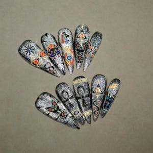 May include: A set of ten long, glittery silver press-on nails. Each nail features a different design, including Egyptian symbols like ankhs, all-seeing eyes, and cat-like figures, in black, red, and gold.