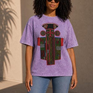K&ouml;nnte beinhalten: Ein violettes T-Shirt in Acid-Wash-Optik mit einem gro&szlig;en, farbenfrohen Grafikdruck einer stilisierten, geometrischen totemartigen Figur mit komplexen Mustern in Rot, Gr&uuml;n, Schwarz und Gelb, kombiniert mit einer klassischen blauen Jeans.