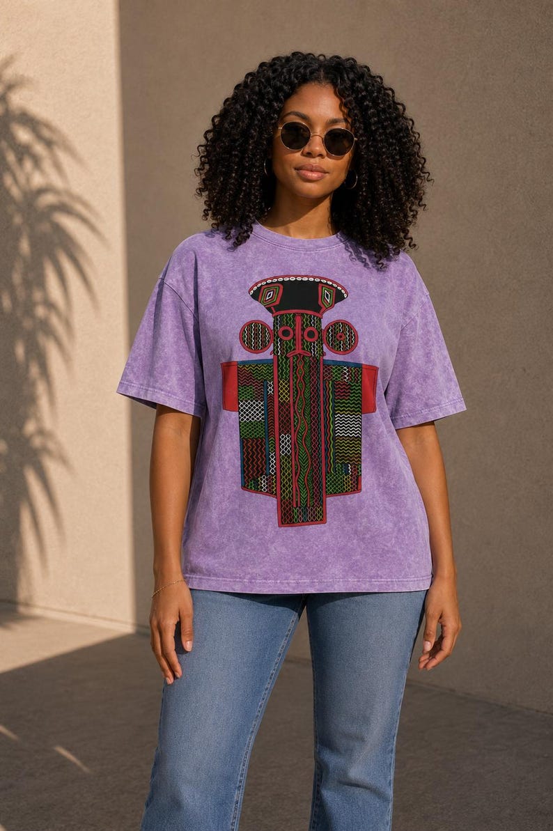 K&ouml;nnte beinhalten: Ein violettes T-Shirt in Acid-Wash-Optik mit einem gro&szlig;en, farbenfrohen Grafikdruck einer stilisierten, geometrischen totemartigen Figur mit komplexen Mustern in Rot, Gr&uuml;n, Schwarz und Gelb, kombiniert mit einer klassischen blauen Jeans.