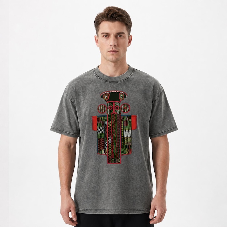 K&ouml;nnte beinhalten: Ein anthrazitfarbenes T-Shirt in Acid-Wash-Optik mit einem zentralen Grafikdesign. Der Druck zeigt eine stilisierte, symmetrische Figur mit einem geometrischen Muster in Rot, Gr&uuml;n und Schwarz, akzentuiert durch wei&szlig;e Punkte und eine rote Basis.