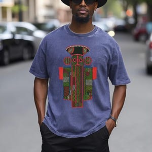 K&ouml;nnte beinhalten: Ein blaues kurz&auml;rmeliges T-Shirt mit einem zentralen Grafikdruck mit geometrischen Mustern in Rot, Gr&uuml;n und Gelb. Das Design zeigt eine stilisierte Figur mit kreisf&ouml;rmigen Elementen und einem flachen Oberteil auf verwaschenem blauem Stoff.