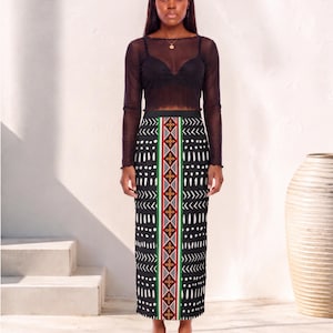 May include: A long black skirt featuring a central vertical panel with a geometric pattern in red, green, and gold, flanked by white dash and chevron motifs on a black background, paired with a black long-sleeved sheer top and white sandals.