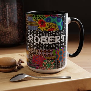 Może przedstawiać: Ceramiczny kubek z czarnym uchwytem i wnętrzem, ozdobiony żywym wzorem patchworkowym składającym się z różnych kolorowych motywów oraz imieniem ROBERT wydrukowanym białymi wielkimi literami na środku zewnętrznej powierzchni.