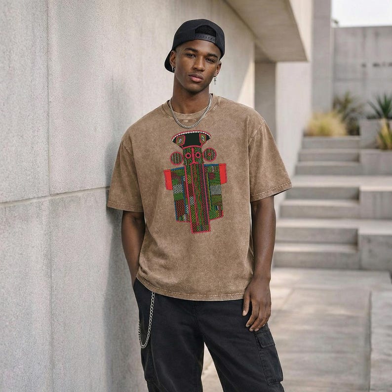K&ouml;nnte beinhalten: Ein braunes T-Shirt im Acid-Wash-Look mit einer zentralen Grafik einer stilisierten Figur mit roten, gr&uuml;nen und schwarzen geometrischen Mustern. Dazu werden eine schwarze Baseballkappe, eine silberne Kette und eine schwarze Cargohose getragen.