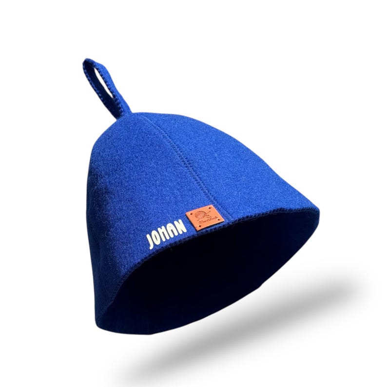 May include: A royal blue sauna hat with a loop at the top. The hat has the word "JOHAN" printed in white on the side, along with a small brown leather patch. The hat is designed to protect the head from heat.