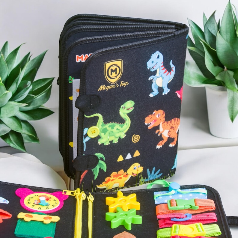 Op de afbeelding: Een zwart stoffen activiteitenboek voor peuters met kleurrijke dinosaurusillustraties. Het boek heeft interactieve elementen zoals ritsen, gespen en tandwielen. De omslag toont de tekst "Megan's Toys".