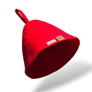 May include: A vibrant red sauna hat with a loop for hanging. The hat features the name "JOHAN" in white lettering and a small brown leather patch with a logo. This hat is designed for use in saunas.