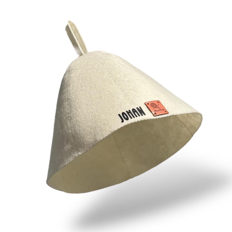 May include: A light beige sauna hat with a conical shape and a small loop at the top. The word "JOHAN" is printed in black on the side, with a small brown leather tag. The hat is designed to protect the head from heat.