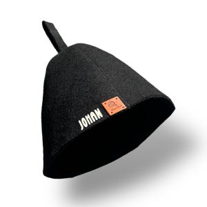 May include: A black sauna hat with a small loop at the top. The word "JOHAN" is printed in white on the side. A small brown leather patch with a logo is also visible.
