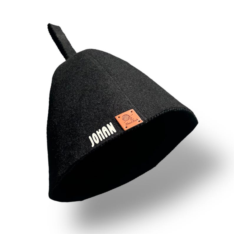 May include: A black sauna hat with a small loop at the top. The word "JOHAN" is printed in white on the side. A small brown leather patch with a logo is also visible.