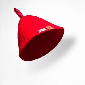 Può includere: Un cappello da sauna rosso vivo con un passante e il nome "JOHAN" a lettere bianche. È visibile anche una piccola etichetta quadrata marrone. Il cappello è realizzato in un materiale morbido ed è progettato per proteggere la testa dal calore.