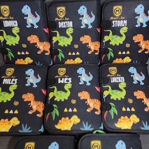 Pode incluir: Uma cole&ccedil;&atilde;o de estojos retangulares pretos com um padr&atilde;o repetitivo de dinossauros coloridos em azul, verde e laranja. Cada estojo exibe um log&oacute;tipo de escudo amarelo com o texto Megan's Toys e um nome &uacute;nico: Ludovico, Daxton, Storm, Miles, Wes ou Lorenzo.