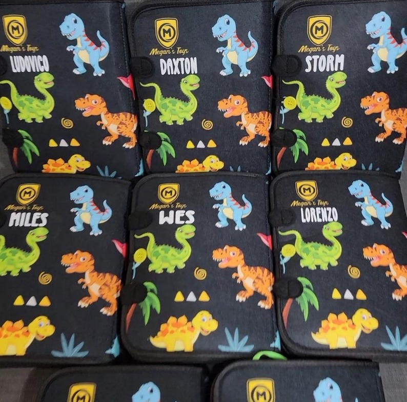 Pode incluir: Uma cole&ccedil;&atilde;o de estojos retangulares pretos com um padr&atilde;o repetitivo de dinossauros coloridos em azul, verde e laranja. Cada estojo exibe um log&oacute;tipo de escudo amarelo com o texto Megan's Toys e um nome &uacute;nico: Ludovico, Daxton, Storm, Miles, Wes ou Lorenzo.