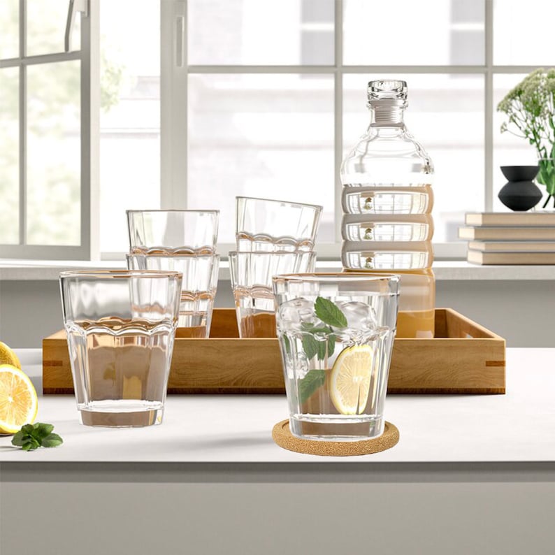 May include: A set of six clear glass tumblers with a textured design, a clear glass bottle, and a cork coaster. The glasses are arranged on a wooden tray. A lemon wedge and a sprig of mint are in one of the glasses.
