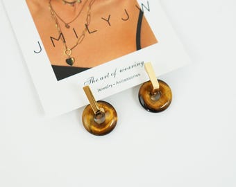 May include: Gold-coloured earrings featuring a circular tiger's eye stone design. Each earring has a rectangular gold bar at the top. The earrings are presented on a white surface with a card that reads "The art of wearing Jewelry Accessories".