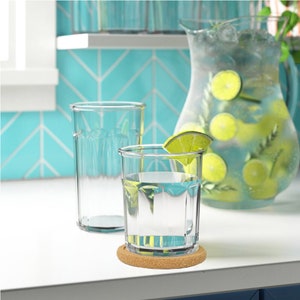 May include: Two clear glass tumblers with a ribbed design, one filled with water and a lime wedge, sitting on a cork coaster. A glass jug filled with water and lime slices is in the background.