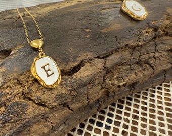 May include: Two gold necklaces with white enamel pendants featuring the letters "E" and "G". The necklaces are displayed on a wooden surface.