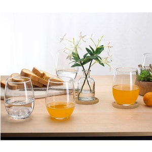 May include: Four clear glass wine glasses with two filled with orange juice and one filled with iced water. The glasses are on wooden coasters on a light wood table. A small glass vase with white flowers is in the centre of the table.