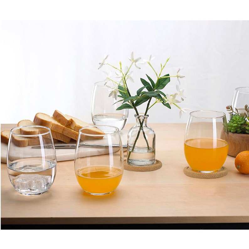 May include: Four clear glass wine glasses with two filled with orange juice and one filled with iced water. The glasses are on wooden coasters on a light wood table. A small glass vase with white flowers is in the centre of the table.