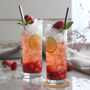 May include: Two tall glasses filled with ice, strawberry slices, and a pink drink.  Lime wedges and fresh mint garnish the drinks.  The glasses are on a light brown surface with a coaster under one glass.