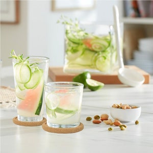 May include: Two clear glass tumblers with sliced cucumbers and watermelon in them, sitting on cork coasters. A small white bowl with pistachios is on a white table.