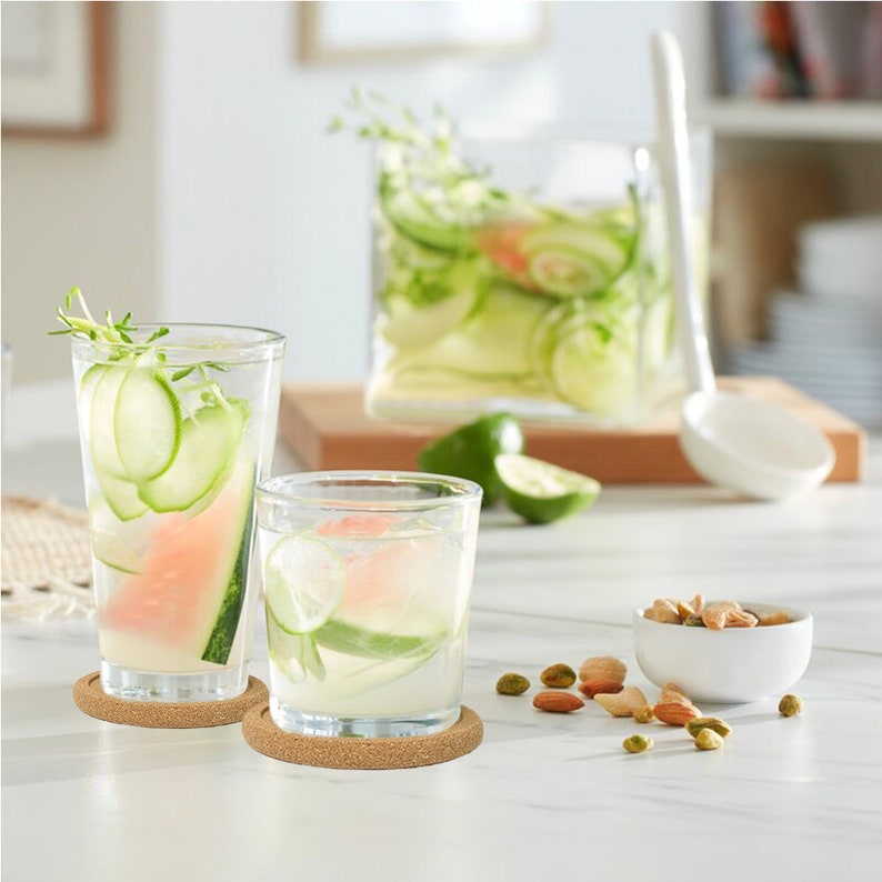 May include: Two clear glass tumblers with sliced cucumbers and watermelon in them, sitting on cork coasters. A small white bowl with pistachios is on a white table.