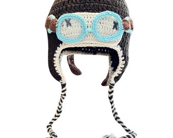 May include: A handmade crochet aviator hat in brown, cream, and blue. The hat features a round crown, ear flaps, and a pair of goggles with blue frames and black cross details. The hat has long, striped ties with tassels.