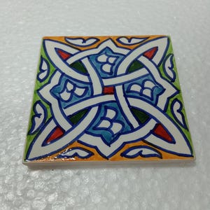 May include: A square ceramic tile with a colorful geometric pattern. The tile features a white background with intricate blue, green, red, and orange designs.