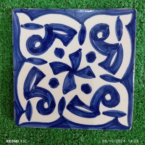 May include: A white ceramic tile with a blue and white geometric pattern. The pattern features a central flower shape with four petals and a circle in the center. The tile is surrounded by a border of blue and white swirls.