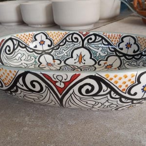 May include: An oval ceramic bowl with a detailed, hand-painted design. The bowl features a white base with a black, red, orange, and blue floral pattern. The rim is a light green color. Other white bowls are in the background.