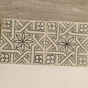 May include: Two square ceramic tiles with a geometric design. The tiles are off-white with black outlines and a pale green background. The design includes floral and geometric shapes.