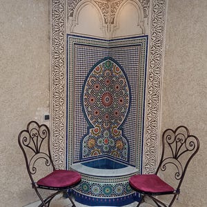 Op de afbeelding: Twee sierlijke ijzeren stoelen met rode kussens staan tegenover een betegelde fontein met een blauw en wit moza&iuml;ekontwerp. De fontein is geplaatst tegen een muur met ingewikkelde witte houtsnijwerk. Een ronde tafel met een kleurrijk moza&iuml;ekblad staat voor de stoelen.