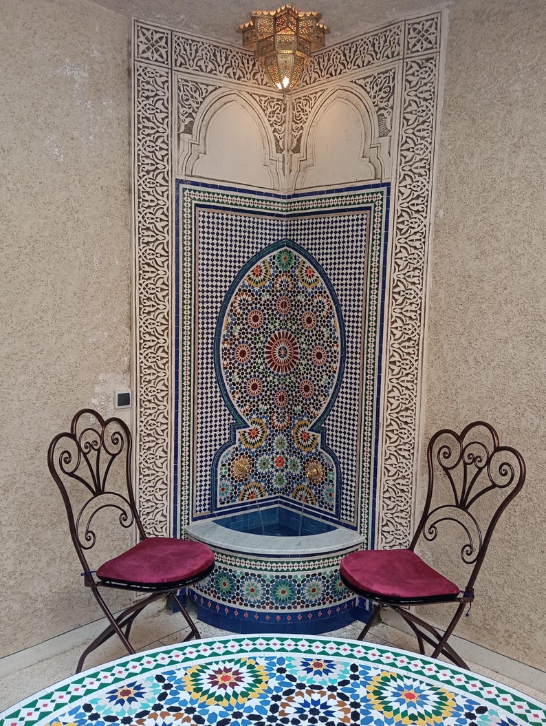 Op de afbeelding: Twee sierlijke ijzeren stoelen met rode kussens staan tegenover een betegelde fontein met een blauw en wit moza&iuml;ekontwerp. De fontein is geplaatst tegen een muur met ingewikkelde witte houtsnijwerk. Een ronde tafel met een kleurrijk moza&iuml;ekblad staat voor de stoelen.