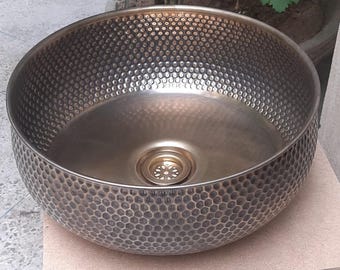 May include: A hammered metal vessel sink with a bronze finish. The sink has a round shape with a textured, dimpled surface. The interior is smooth with a central drain. The sink is displayed on a light-colored wooden surface.