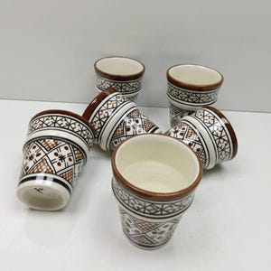 Könnte beinhalten: Ein Set aus sechs Keramikbechern mit weißem Grund, braunem Rand und komplizierten geometrischen Mustern in Schwarz und Braun. Die Becher sind auf einer weißen Oberfläche angeordnet.