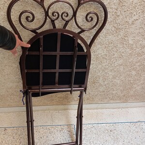 Op de afbeelding: Een bruine metalen vouwstoel met een zwarte stoffen zitting en een decoratieve, gerolde rugleuning.