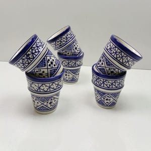 Könnte beinhalten: Sechs kleine, gestapelte Keramikbecher mit blau-weißem geometrischen Muster. Die Becher haben eine ausgestellte Form mit blauem Rand und detailliertem Design. Sie sind auf einer weißen Oberfläche angeordnet.