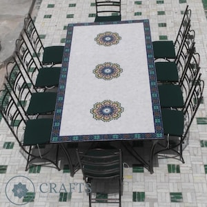 Peut inclure: Un ensemble de salle à manger d'extérieur avec une table rectangulaire ornée d'un dessus en mosaïque blanc avec trois motifs floraux. Il est entouré de chaises rembourrées vert foncé et de cadres en métal noir ornés. Idéal pour le jardin.