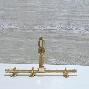 Könnte beinhalten: Ein goldfarbener Metall-Kleiderständer mit dekorativem Design. Der Ständer hat einen vertikalen Griff mit seilartigem Design und eine horizontale Stange mit mehreren Haken. Der Ständer ist für die Wandmontage konzipiert.