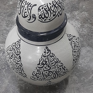 May include: A white ceramic jar with a lid, adorned with black Arabic calligraphy. The jar has a bulbous body and a lid with a black knob. The calligraphy is a prominent design element, adding a cultural touch to the piece.