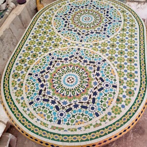 Könnte beinhalten: Ein ovaler Mosaiktisch mit farbenfrohem geometrischen Design. Die Tischplatte weist komplizierte Muster in Blau-, Grün-, Gelb- und Weißtönen auf. Der Rand des Tisches hat ein sich wiederholendes Blumenmotiv. Der Tisch ist etwa 152 cm lang und 76 cm breit.