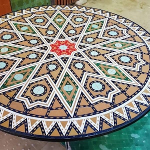 May include: A round table with an intricate mosaic design. The mosaic features a starburst pattern with geometric shapes in shades of tan, green, red, and blue. The table's edge is dark, and the overall design is symmetrical.