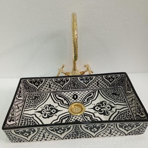 May include: A black and white ceramic sink with a decorative pattern. The sink has a gold-coloured drain and a gold-coloured faucet.