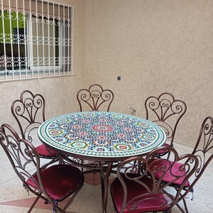 Op de afbeelding: Een ronde tafel met moza&iuml;ektegels en een gesmeed ijzeren frame en zes bijpassende stoelen. De stoelen hebben een gerolde vormgeving en bordeauxrode fluwelen kussens. De tafel staat op een betegelde vloer.
