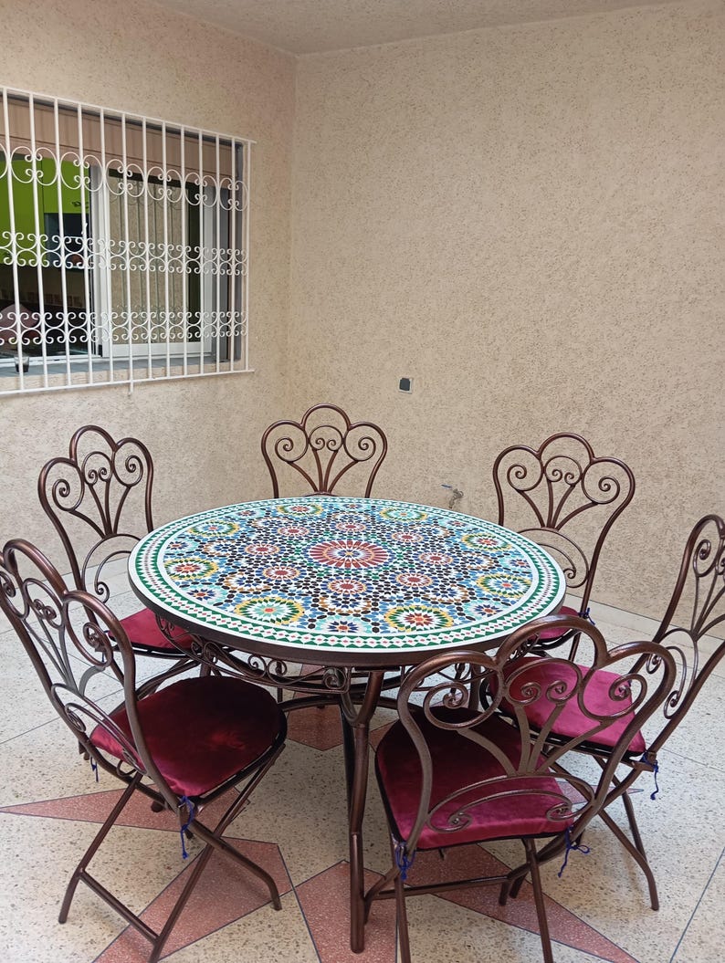 Op de afbeelding: Een ronde tafel met moza&iuml;ektegels en een gesmeed ijzeren frame en zes bijpassende stoelen. De stoelen hebben een gerolde vormgeving en bordeauxrode fluwelen kussens. De tafel staat op een betegelde vloer.