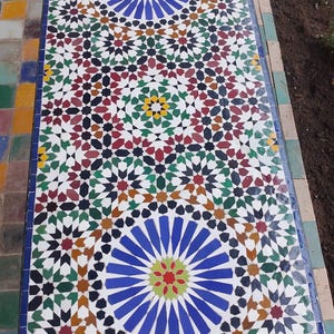May include: A rectangular mosaic table with an intricate geometric design. The pattern incorporates star shapes in blue, red, green, black, and white. The table has a blue border. The table's design is likely for outdoor use.