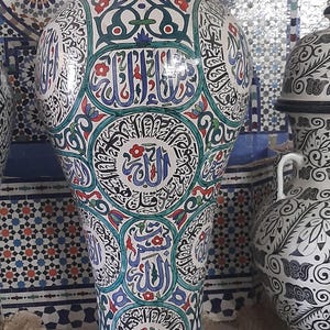 May include: A white ceramic vase with intricate blue, green, and red Arabic calligraphy and floral designs. The vase has a rounded top and a narrow base.
