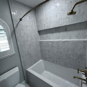 May include: A modern bathroom with a white bathtub, toilet, and gray tiled walls. The shower features a gold-colored showerhead and curved rod. The room has a window with white shutters.