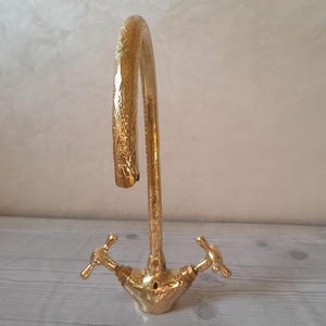 May include: A gold-plated kitchen faucet with a decorative design and two handles. The faucet has a high arc spout and is designed for a single-hole installation.